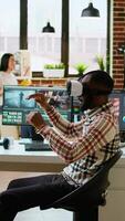 Vertical African american editor uses virtual reality glasses to do color grading, working with high tech vr headset and dual monitors at his home office. Filmmaker editing clips. Camera A. video