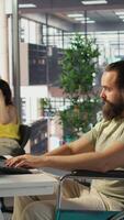 Vertical IT worker with disability sitting in wheelchair in office working at computer desk, focused on testing programs. Software engineer with paraplegia in inclusive workplace checking code on PC, camera B video