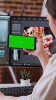 Vertical Asian female editor working from home for freelance work with green screen, using smartphone and dual monitors for mixing software. Creative workspace equipped with tech tools. Camera B. video