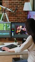 Vertical Asian woman editing a music in her living room studio, multitasking between timeline editing and color grading. Professional workspace features tech tools for multimedia creation. Camera A. video
