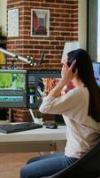 Vertical Asian filmmaker woman putting headphones on and working on production at a home office, mixing clips. Office setup with a software perfect for color grading in post production. Camera A. video