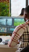 Vertical Male editor works from modern home office in the living room, multitasking with a dual monitor setup and editing software. Finishing post production tasks for media production. Camera B. video