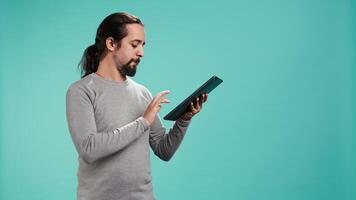 Portrait of upbeat person mindlessly scrolling on tablet screen, isolated over studio background. Cheerful man passing time on social media on portable device, enjoying leisure time, camera B video
