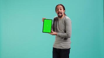 Happy man holding green screen tablet, doing recommendation. Cheerful person presenting mockup device, showing positive feedback, isolated over studio background, camera A video