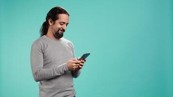 Happy man texting best friend using smartphone, reading messages. Relaxed person having fun while chatting with mate on mobile phone, isolated over blue studio background, camera B video