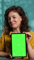 Vertical Jolly woman filming tech tutorial on how to repair damaged mockup tablet. Joyous tech professional in home studio teaching subscribers to install operating system on isolated screen device, camera A video