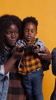 Vertical Cheerful african american dad playing games with his son on console, using controller to have fun against yellow background. Happy positive young family enjoying their leisure time. Camera B. video