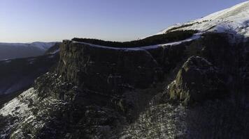 The slope of the high rock massif covered with pine trees on blue clear sky background. Shot. Aerial of the forested mountain slope and snowy peak. photo