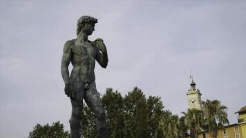 Statue of David in park. Action. Beautiful manly statue of David stands in park on background of sky. Nude statue of David is made of dark stone and stands on pedestal in park photo