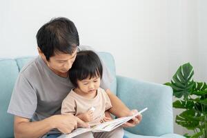 Happy Asian father relax and read book with baby time together at home. parent sit on sofa with daughter and reading a story. learn development, childcare, laughing, education, storytelling, practice. photo
