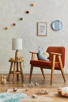 Stylish composition of cozy scandinavian child's room interior with mock up poster frame, red armchair, elegant lamp, plush toys and hanging decorations. Creative wall, carpet on the floor. Template. photo