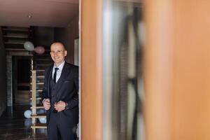 A man in a suit is standing in front of a door. He is smiling and looking at the camera photo