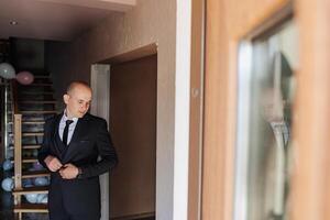 A man in a suit is getting ready to leave the house. He is wearing a tie and adjusting his jacket. The scene is set in a home, with a staircase visible in the background photo