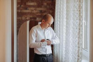 A man in a white shirt is getting dressed in front of a window. He is adjusting his shirt and looking out the window photo