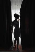 A woman in a white gown stands in front of a curtain. The curtain is drawn, and the woman is partially hidden behind it. The image has a mysterious and intimate mood photo
