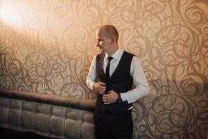 A man in a suit and vest stands in front of a wall with a patterned design. He is adjusting his tie and he is in a professional setting photo