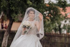 A woman in a white dress is holding a bouquet of flowers and is wearing a veil. She is smiling and she is happy photo