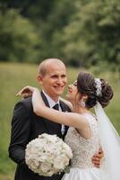 A bride and groom are hugging each other in a field. The bride is wearing a white dress and a veil. They are both smiling and seem happy photo