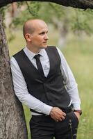 A man in a black vest and white shirt stands next to a tree. He is wearing a black tie and has his hands in his pockets. Concept of formality and elegance photo