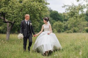 A bride and groom are walking through a field of grass. The bride is wearing a white dress and the groom is wearing a suit. They are holding hands and appear to be happy and in love photo