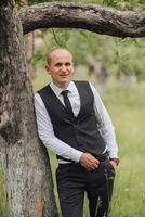 A man in a black vest and tie stands next to a tree. He is smiling and looking at the camera photo