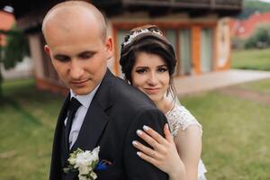 A bride and groom are posing for a picture in front of a house. The bride is wearing a white dress and the groom is wearing a black suit. They are both smiling and seem happy photo