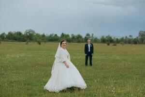 A bride and groom are standing in a field, with the bride wearing a white dress photo