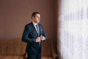 A man in a suit is standing in front of a window, adjusting his tie photo