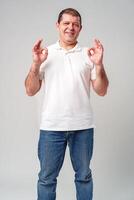 A man in a white shirt poses with a cheerful expression showing OK hand gestures in a well-lit indoor setting photo