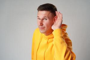 Portrait of young casual man with hand on ear who overhears on gray background photo
