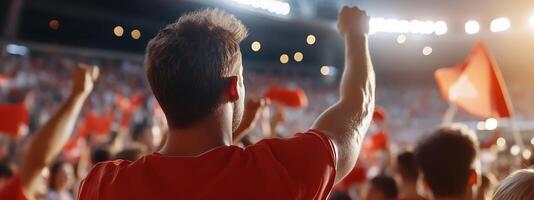 Cheering crowd in red attire raises fists during sporting event at night under bright stadium lights photo