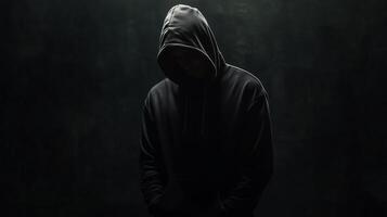 A solitary figure in a dark hoodie stands quietly against a shadowy backdrop, evoking a sense of mystery and introspection in a low-lit environment photo