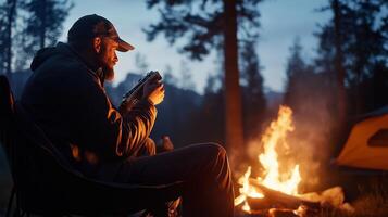 A man enjoys a quiet evening by the campfire in a forested area during twilight, surrounded by tall trees and nature photo