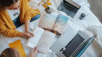 Two individuals studying together indoors, surrounded by books, laptops, and notebooks on a cozy white bed with natural lighting photo