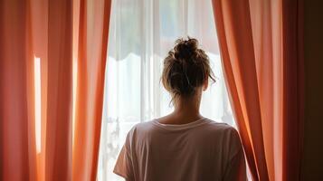 A person gazes out a bright window through sheer curtains in a cozy indoor space during the daytime photo