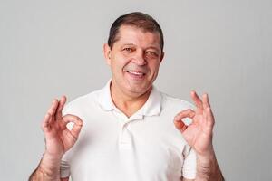 A man in a white shirt poses with a cheerful expression showing OK hand gestures in a well-lit indoor setting photo