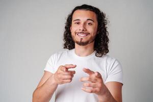 A smiling young man with curly hair playfully pointing directly at the viewer in a light-colored setting photo