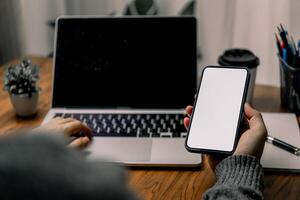 Person using a smartphone while sitting at a desk with a laptop, suggesting multitasking or mobile communication. The screen of the smartphone is blank. photo