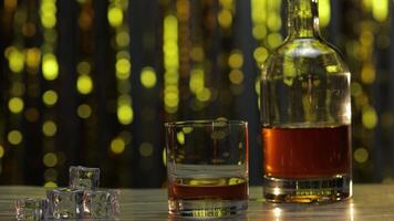 Barman adds ice cubes into glass with golden whiskey, cognac or brandy on table. Shiny background video