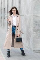 A young pretty girl in a raincoat and jeans walks around the city near a tall building, with a handbag in hand photo