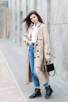 A young pretty girl in a raincoat and jeans walks around the city near a tall building, with a handbag in hand photo