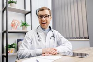 Portrait of young doctor wearing glasses and white uniform with stethoscope, speaking and consulting patient online, looking at camera, making call, sitting at table in office photo