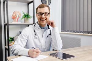 Portrait of young doctor wearing glasses and white uniform with stethoscope, speaking and consulting patient online, looking at camera, making call, sitting at table in office photo
