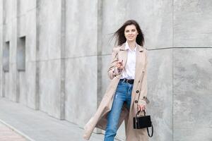 A young pretty girl in a raincoat and jeans walks around the city near a tall building, with a handbag in hand photo