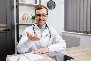 Portrait of young doctor wearing glasses and white uniform with stethoscope, speaking and consulting patient online, looking at camera, making call, sitting at table in office photo