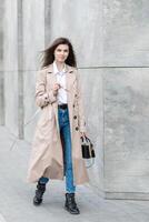 A young pretty girl in a raincoat and jeans walks around the city near a tall building, with a handbag in hand photo