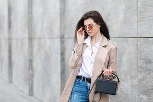 A young pretty girl in a raincoat and jeans walks around the city near a tall building, with a handbag in hand photo