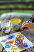 Child dipping paintbrush in colorful jar creating artwork outdoors photo