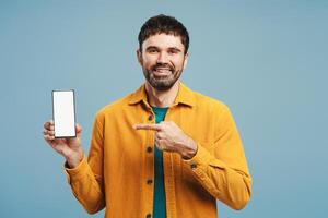 Bearded, middle aged man holding mobile phone with blank screen, pointing finger to mockup photo