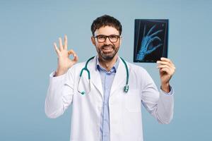 Smiling professional doctor attractive man wearing white coat, holding X-ray picture showing ok sign photo